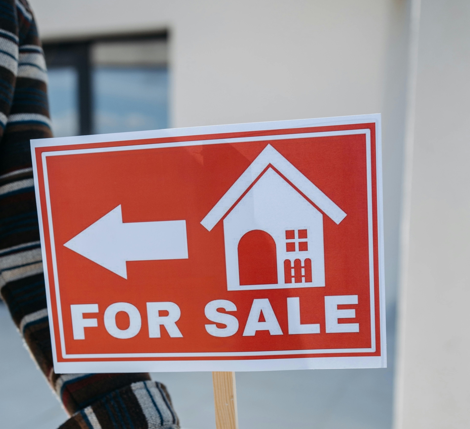 Close-up of a hand holding a 'For Sale' real estate sign with a house icon outdoors.